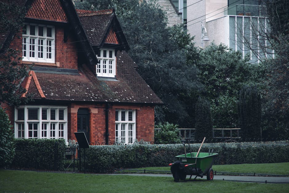 A red brick house with a steeply pitched roof covered in dark tiles, featuring white-framed sash windows and a small, gabled dormer. The front garden is neatly maintained, with a low hedge bordering a